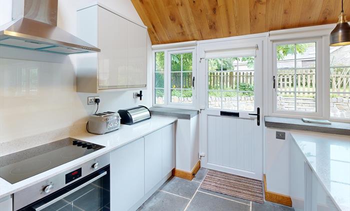 A bright, contemporary kitchen featuring white cabinets, quartz countertops, an electric hob, and a door opening onto a garden with stone walls and fe